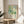 May include: Framed poster with abstract green and blue watercolor design featuring circular dots and organic shapes, displayed on round wooden side table next to beige upholstered chair. Natural sunlight creates soft shadows on white wall. Plant leaves visible in corner. Poster shows Japanese text and "JAPAN 1957" at bottom in minimalist typography.