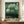 May include: Framed poster with green background featuring stylized trees, polka dots, and a small house in artistic style reminiscent of Kusama's work. The print displays "YAYOI KUSAMA" and "KYOTO" text at bottom with Japanese characters. Poster sits on wooden sideboard next to white ceramic vase and stacked books in modern interior with white walls and trim.