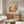 May include: Modern living room with wooden sideboard displaying framed art print featuring abstract botanical design in warm orange, coral, and cream tones with stylized flowers and foliage. Scene includes white curved chair, glass oval coffee table, floor lamp, and neutral wall backdrop with natural lighting.