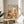 May include: Framed art print displayed on wooden round side table next to beige upholstered chair. Poster features warm orange, cream and brown botanical illustration with stylized mushrooms, leaves and organic shapes in mid-century modern style. Japanese characters visible at top along with "JAPAN 1987" text. Natural lighting creates soft shadows across cream wall background with plant leaves visible in corner.