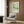 May include: Framed poster with abstract green and yellow botanical design featuring dots and organic shapes, displayed on wood paneled wall above beige upholstered armchair with geometric patterned pillow. Modern reading corner with brass floor lamp, dark round side table with open book, and white curtains. Warm natural lighting on wooden floors creates cozy contemporary interior setting.