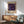 May include: Framed Yayoi Kusama exhibition poster displayed on white wall above wooden sideboard with mint green mid-century armchair beside it. Poster features colorful floral and circular patterns on dark purple background with cream matting and natural wood frame. Table lamp and round coffee table visible in sunlit modern interior room setting.