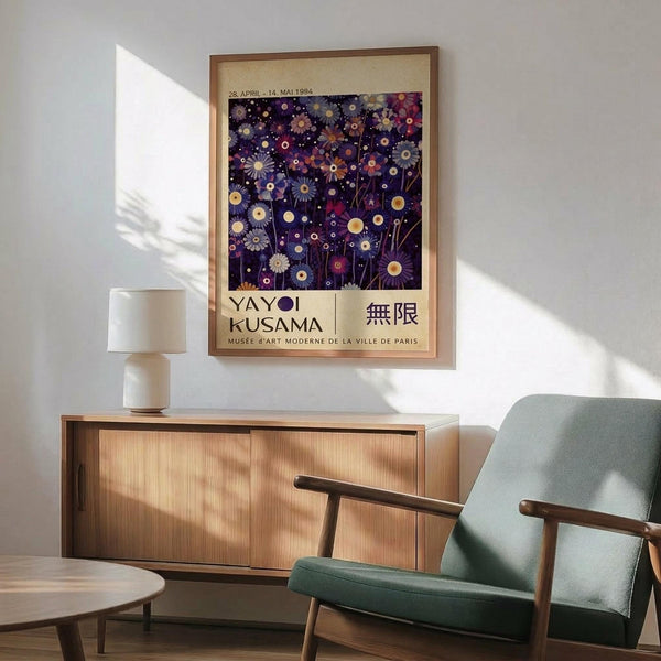 May include: Framed Yayoi Kusama exhibition poster displayed on white wall above wooden sideboard with mint green mid-century armchair beside it. Poster features colorful floral and circular patterns on dark purple background with cream matting and natural wood frame. Table lamp and round coffee table visible in sunlit modern interior room setting.