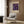 May include: Beige framed poster with purple background featuring colorful floral and circular patterns in blue, pink, and gold tones, displayed on wood paneled wall above light gray upholstered armchair with geometric patterned pillow, next to brass floor lamp and dark round side table with open book