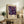 May include: Framed poster with purple background featuring colorful abstract flowers and circles in pink, orange, and yellow tones, displayed on rustic wooden sideboard next to fern plant, ceramic vase, and books in neutral-toned living room setting with beige chair visible.