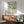 May include: Framed abstract art print with organic green, teal, and yellow shapes and dots on white background, displayed above mid-century modern wooden sideboard with white ceramic lamp. Light green upholstered armchair with wooden arms sits nearby in bright, minimalist living room with white walls and natural sunlight casting shadows.