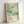 May include: Framed art print leaning against white wall featuring abstract botanical design in soft greens, blues, and yellows with polka dots and organic plant forms. Text reads "YAYOI KUSAMA" with Japanese characters and "JAPAN 1987" in minimal typography. Light wooden frame with natural finish, photographed in bright natural lighting showing subtle shadows.
