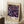 May include: Framed art print leaning against white wall corner on fluffy white rug. Features purple night sky filled with colorful daisy-like flowers in blues, purples, pinks, and yellows with white dots resembling stars. Text shows dates "28. APRIL - 14. MAI 1984" and "YAYOI KUSAMA MUSÉE D'ART MODERNE DE LA VILLE DE PARIS" with Japanese characters. Natural wood frame, cream matting. Small wooden stool with white coffee cup visible on left side.