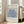 May include: Framed vintage-style exhibition poster featuring blue coral-like botanical cutout shapes on cream background, displayed on light wooden floor against white wall next to beige upholstered sofa with pillows. Natural sunlight streams through white-framed windows creating soft shadows. The artwork shows organic flowing forms in various shades of blue with black text above and below the design.