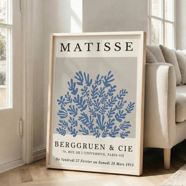 May include: Framed vintage-style exhibition poster featuring blue coral-like botanical cutout shapes on cream background, displayed on light wooden floor against white wall next to beige upholstered sofa with pillows. Natural sunlight streams through white-framed windows creating soft shadows. The artwork shows organic flowing forms in various shades of blue with black text above and below the design.