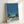 May include: Framed art print leaning against white wall showing retro-style illustration of mid-century modern building with swimming pool in foreground, tall palm trees against bright blue sky, and "PROUD" text on building facade. Light wood frame, contemporary room setting with natural lighting casting soft shadows.