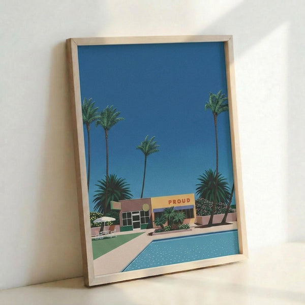 May include: Framed art print leaning against white wall showing retro-style illustration of mid-century modern building with swimming pool in foreground, tall palm trees against bright blue sky, and "PROUD" text on building facade. Light wood frame, contemporary room setting with natural lighting casting soft shadows.