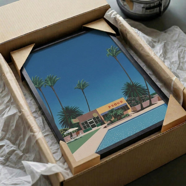 May include: Framed poster print being unboxed from cardboard shipping package with protective wrapping. Artwork shows retro-style illustration of tropical poolside scene with tall palm trees, turquoise swimming pool, and mid-century modern building with "PROUD" signage against bright blue sky. Print appears medium-sized in black frame surrounded by tissue paper and cardboard packaging materials.