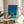 May include: Framed artwork displaying a retro-style illustration of a mid-century modern house with swimming pool under bright blue sky, surrounded by tall palm trees. The poster features vibrant blues, greens, and warm earth tones in a nostalgic aesthetic. Wooden frame sits on round wooden side table next to beige upholstered chair, with plant shadows cast on white wall from natural sunlight.