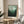 May include: Framed forest landscape poster with green misty trees and pale sky, displayed on round wooden side table next to beige upholstered chair with throw blanket. Natural sunlight streams across neutral walls creating soft shadows. Plant leaves visible in corner.
