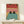 May include: Framed poster print showing two black cats with yellow eyes sitting in a turquoise blue bathtub or pool, set against coral pink tiled bathroom walls. The illustration has a vintage comic book style with bold colors and black outlines. The wooden frame leans against a white wall on a light wood shelf next to a small wrapped gift with twine and dried plant sprig.