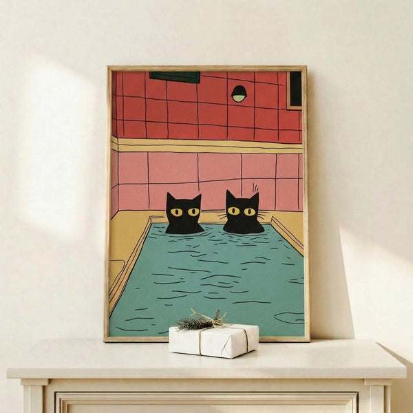 May include: Framed poster print showing two black cats with yellow eyes sitting in a turquoise blue bathtub or pool, set against coral pink tiled bathroom walls. The illustration has a vintage comic book style with bold colors and black outlines. The wooden frame leans against a white wall on a light wood shelf next to a small wrapped gift with twine and dried plant sprig.