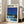 May include: Framed art print leaning against white wall beside beige sofa in bright living room with large windows. Poster features modernist illustration of mid-century home with swimming pool, palm trees against deep blue sky. Clean geometric architecture with glass walls and yellow accents. Print has Japanese text at top and light wood frame.