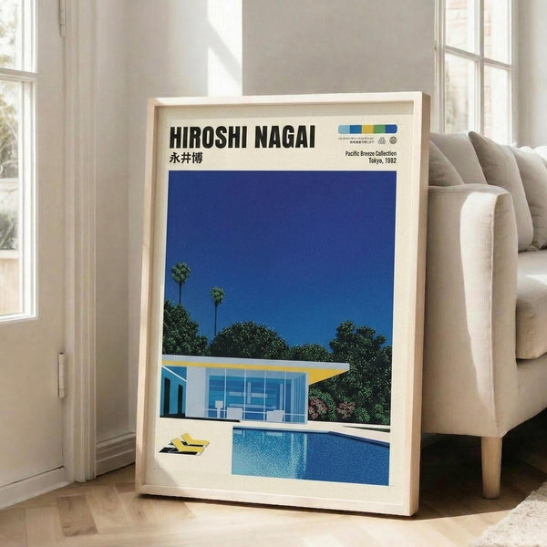 May include: Framed art print leaning against white wall beside beige sofa in bright living room with large windows. Poster features modernist illustration of mid-century home with swimming pool, palm trees against deep blue sky. Clean geometric architecture with glass walls and yellow accents. Print has Japanese text at top and light wood frame.