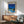 May include: Framed poster showing modern white house with swimming pool under bright blue sky with palm trees, displayed above wooden sideboard with white ceramic lamp in mid-century modern living room with sage green upholstered chair and round coffee table
