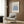 May include: Framed Matisse exhibition poster with blue botanical cutout design hanging on wood-paneled wall above beige upholstered chair with geometric pillow. Modern floor lamp with brass finish and small round side table with open book visible. Neutral toned room setting with natural lighting from window.
