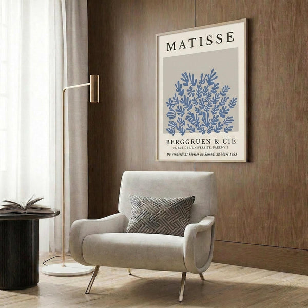 May include: Framed Matisse exhibition poster with blue botanical cutout design hanging on wood-paneled wall above beige upholstered chair with geometric pillow. Modern floor lamp with brass finish and small round side table with open book visible. Neutral toned room setting with natural lighting from window.