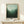 May include: Framed forest landscape poster in misty green tones leaning against white wall on light wood console table, with small wrapped white gift box beside it. The artwork shows silhouetted evergreen trees emerging from fog, transitioning from pale mint green sky to deep forest green at bottom. Natural wood frame surrounds the print which includes exhibition text at bottom.