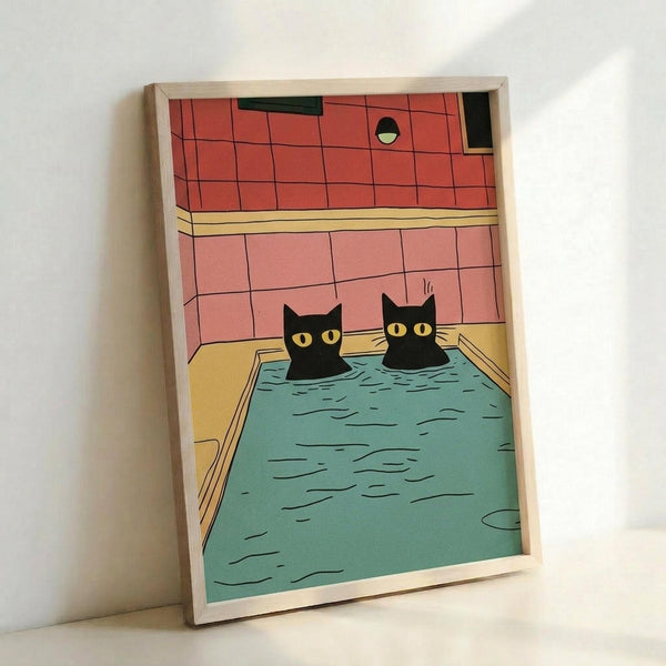 Two black cats with yellow eyes relaxing in a turquoise bathtub, illustrated in a minimalist cartoon style. The bathroom scene features coral pink tile walls and warm yellow tub edges. The artwork is displayed in a light wood frame leaning against a white wall, showing the print's actual colors and scale.