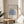 May include: Framed Matisse exhibition poster with blue coral-like cutout design on beige background, displayed on round wooden side table next to beige upholstered chair. Natural lighting creates soft shadows across white walls. Green plant leaves visible in corner. Poster shows gallery text "Berggruen & Cie" with Paris address and 1953 exhibition dates in thin wooden frame.