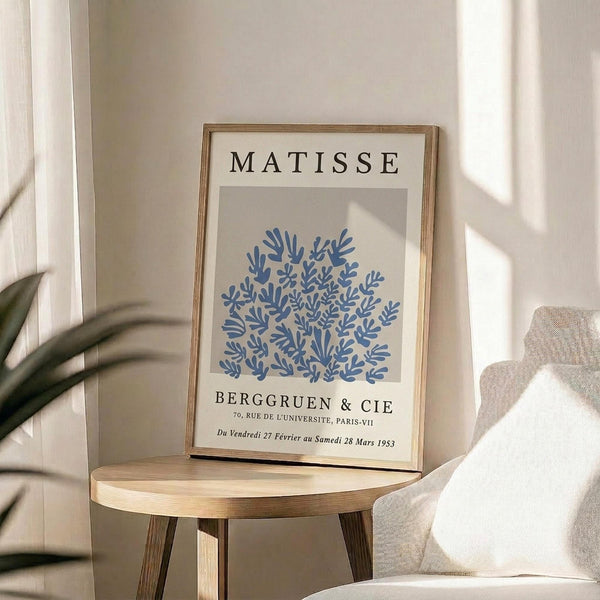 May include: Framed Matisse exhibition poster with blue coral-like cutout design on beige background, displayed on round wooden side table next to beige upholstered chair. Natural lighting creates soft shadows across white walls. Green plant leaves visible in corner. Poster shows gallery text "Berggruen & Cie" with Paris address and 1953 exhibition dates in thin wooden frame.