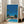 May include: Framed poster print leaning against beige sofa in bright living room with white walls and large windows. Artwork shows retro-style illustration of mid-century modern house with swimming pool, tall palm trees, and vibrant blue sky. Orange "PROUD" sign visible on pink building. Tropical poolside scene rendered in flat, stylized aesthetic with bold colors including turquoise pool water and lush green palm fronds.