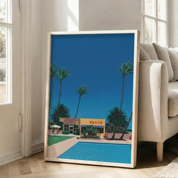 May include: Framed poster print leaning against beige sofa in bright living room with white walls and large windows. Artwork shows retro-style illustration of mid-century modern house with swimming pool, tall palm trees, and vibrant blue sky. Orange "PROUD" sign visible on pink building. Tropical poolside scene rendered in flat, stylized aesthetic with bold colors including turquoise pool water and lush green palm fronds.
