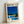 May include: Framed poster print leaning against white wall showing modernist poolside scene with glass house, bright blue swimming pool, palm trees, and deep blue sky. Light wood frame surrounds white-bordered print with "HIROSHI NAGAI" text at top. Clean minimalist aesthetic with vibrant blues, yellows, and greens depicting sunny California-style architecture and landscape.