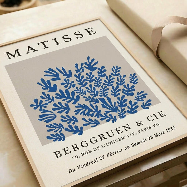 May include: Framed art print featuring blue coral-like botanical forms on beige background. White matted frame sits on neutral surface next to rolled cream paper and ribbon. Gallery-style poster with "MATISSE" at top, "BERGGRUEN & CIE" below artwork, includes Paris address and 1953 exhibition dates in French. Minimalist styling with warm lighting creates elegant display scene.
