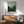 May include: Framed forest landscape poster with misty green pine trees and pale sky, displayed above wooden sideboard with table lamp in modern living room setting. Mid-century style green upholstered chair with wooden arms sits nearby, creating cozy interior scene with natural sunlight streaming across white walls.