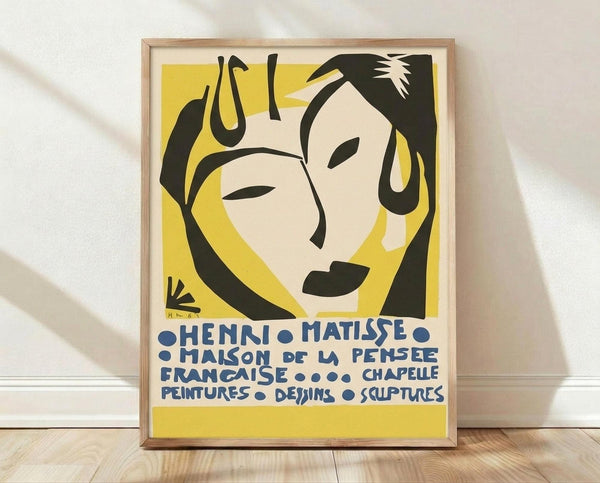 May include: Framed vintage exhibition poster featuring abstract portrait of woman with black hair against yellow background, displayed on white textured wall above light wood flooring. French text in blue letters advertises Matisse artwork including paintings, drawings, and sculptures. Natural wood frame complements minimal interior setting with soft shadows.