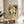 May include: Framed vintage French exhibition poster with stylized abstract face illustration in yellow, black and cream tones, displayed on round wooden side table next to beige upholstered chair. Natural sunlight streams across white wall creating geometric shadows. Green plant leaves visible in corner of minimalist modern living room setting.