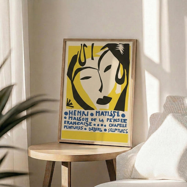 May include: Framed vintage French exhibition poster with stylized abstract face illustration in yellow, black and cream tones, displayed on round wooden side table next to beige upholstered chair. Natural sunlight streams across white wall creating geometric shadows. Green plant leaves visible in corner of minimalist modern living room setting.