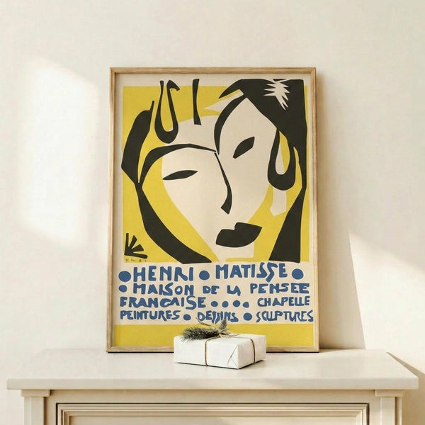 May include: Framed Henri Matisse exhibition poster with yellow background featuring abstract black silhouettes of two faces in profile, blue text in French mentioning paintings, drawings and sculptures. Wooden frame displayed on light wood shelf next to small wrapped white gift box with natural twine and dried plant sprig against cream wall.