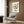 May include: Modern living room with beige upholstered armchair and geometric patterned pillow. Framed poster on wood-paneled wall shows vinyl record design with red center, black tonearm, and text spiraling around the disc reading "Song Name" and "Artist Name." Golden floor lamp and dark round side table with open book visible. Warm wood tones and neutral color palette throughout the contemporary interior space.