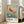 May include: Framed vintage-style Japanese art print featuring an orange and red koi fish swimming among delicate white cherry blossoms against a muted blue-green background. The poster displays "OHARA SHOSON" text and Japanese characters at the bottom with museum exhibition details from 1972. The wooden-framed artwork leans against a white wall next to a beige upholstered chair in a bright, minimalist living room with large windows.