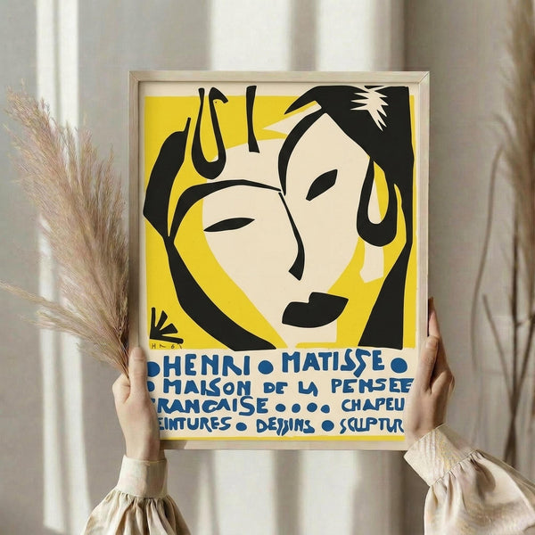 May include: Person in cream ruffled sleeves holding framed Henri Matisse exhibition poster with yellow background featuring abstract black line art face design and blue French text. Dried pampas grass visible beside frame against soft white curtained window backdrop.