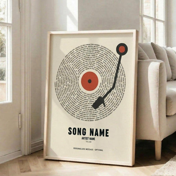 May include: Framed poster with vinyl record design featuring song lyrics arranged in concentric circles around an orange center label, black tonearm detail, and placeholder text for song name, artist name, and year below. Light wood frame leaning against white wall near beige upholstered chair in bright room with large windows.