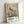 May include: Framed vintage-style exhibition poster leaning against white wall, featuring Japanese artwork of golden-orange koi fish swimming among white cherry blossoms in blue-green water with rain-like streaks. Light wooden frame with white matting. Text shows "OHARA SHOSON" with Japanese characters and museum exhibition details from 1972. Soft natural lighting creates gentle shadows on wall.