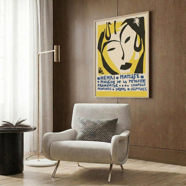 May include: A framed Henri Matisse exhibition poster featuring a stylized woman's face in yellow, black and cream colors with French text below. The poster hangs on a wood-paneled wall above a beige upholstered armchair with geometric patterned pillow. A brass floor lamp and dark side table with open book complete the modern living room setting with natural lighting from a nearby window.