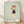 May include: Framed poster print leaning against white wall on light wood floor. The artwork features a stylized cartoon character with reddish-brown bob haircut and bunny ears, wearing a dark dress with arms crossed and mischievous expression, set against pale mint green background. Text below shows "YOSHITOMO NARA" in black letters with Japanese characters and exhibition dates. Natural wood frame with white matting.
