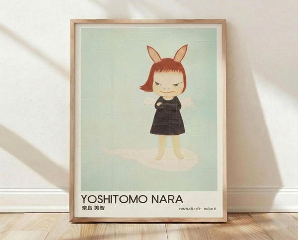 May include: Framed poster print leaning against white wall on light wood floor. The artwork features a stylized cartoon character with reddish-brown bob haircut and bunny ears, wearing a dark dress with arms crossed and mischievous expression, set against pale mint green background. Text below shows "YOSHITOMO NARA" in black letters with Japanese characters and exhibition dates. Natural wood frame with white matting.