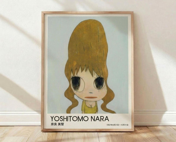 May include: Framed poster print featuring anime-style illustration of young girl with large golden blonde hair in ponytails, oversized dark eyes, and yellow top against pale blue background. Text reads "YOSHITOMO NARA" with Japanese characters and "1992年8月31日〜10月31日" below. Natural wood frame displayed on light wooden floor against white textured wall with diagonal shadows.