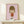 May include: Framed art print featuring a stylized cartoon girl with olive green bob haircut and closed eyes, wearing an orange-red dress, displayed on light wood shelf. Pink background artwork in thin gold frame alongside small white wrapped gift with evergreen sprig. Minimalist nursery or bedroom setting with cream walls.