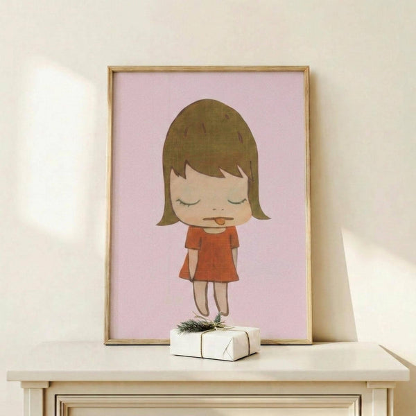 May include: Framed art print featuring a stylized cartoon girl with olive green bob haircut and closed eyes, wearing an orange-red dress, displayed on light wood shelf. Pink background artwork in thin gold frame alongside small white wrapped gift with evergreen sprig. Minimalist nursery or bedroom setting with cream walls.